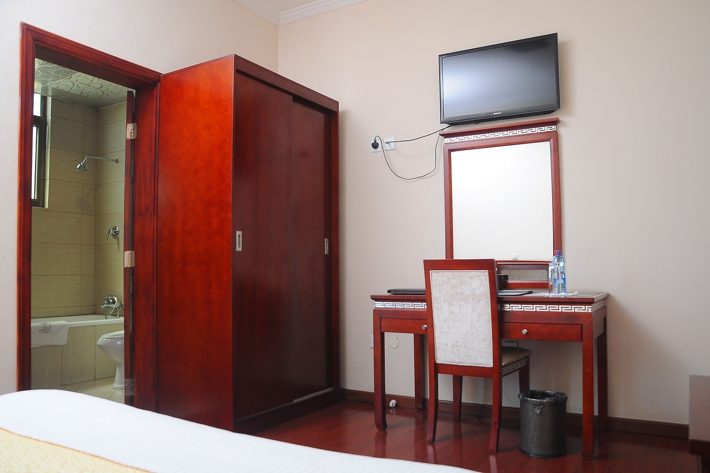 A hotel room with a wooden wardrobe, a desk with a chair, a television mounted above the desk, and a trash bin.
