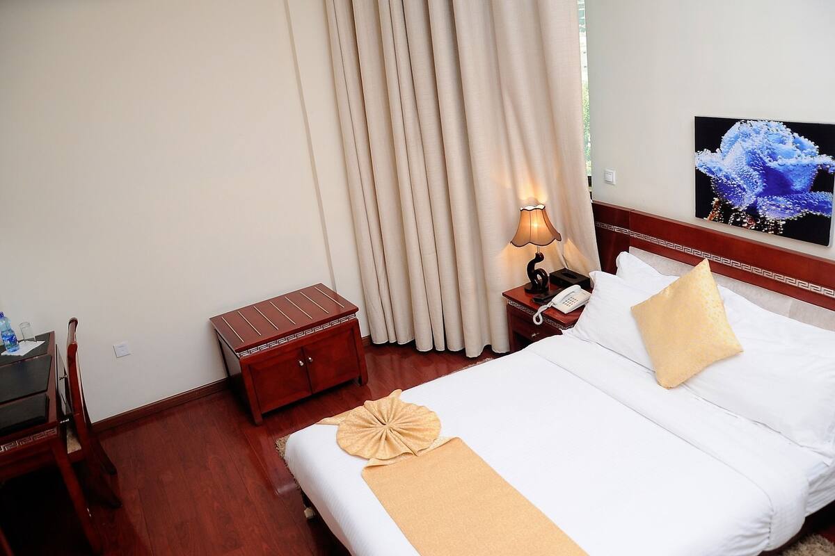 A hotel room with a bed, a nightstand with a lamp, a wooden chest, and a wall-mounted phone.