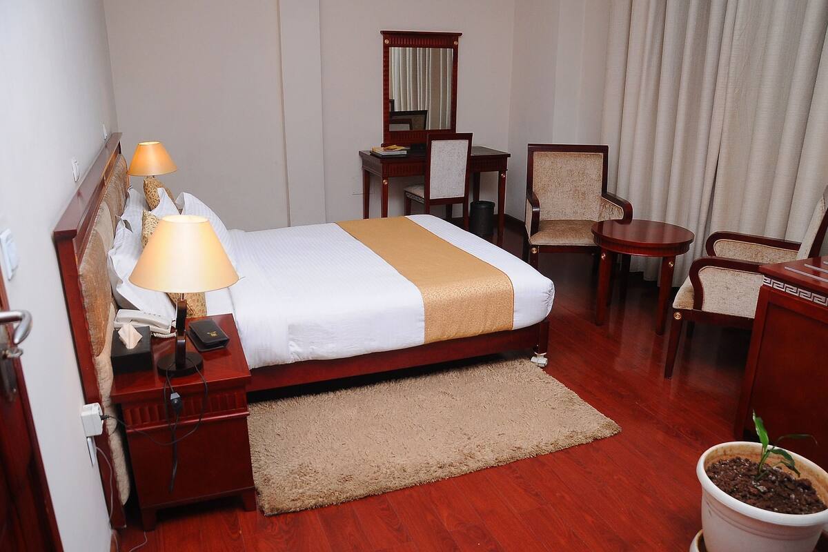A hotel room with a bed, bedside lamps, a desk, a chair, a small table, and a potted plant.