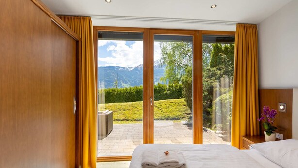 Ein Hotelzimmer mit Bett, einem kleinen Tisch und Blick auf eine gebirgige Landschaft durch große Glasfenster.