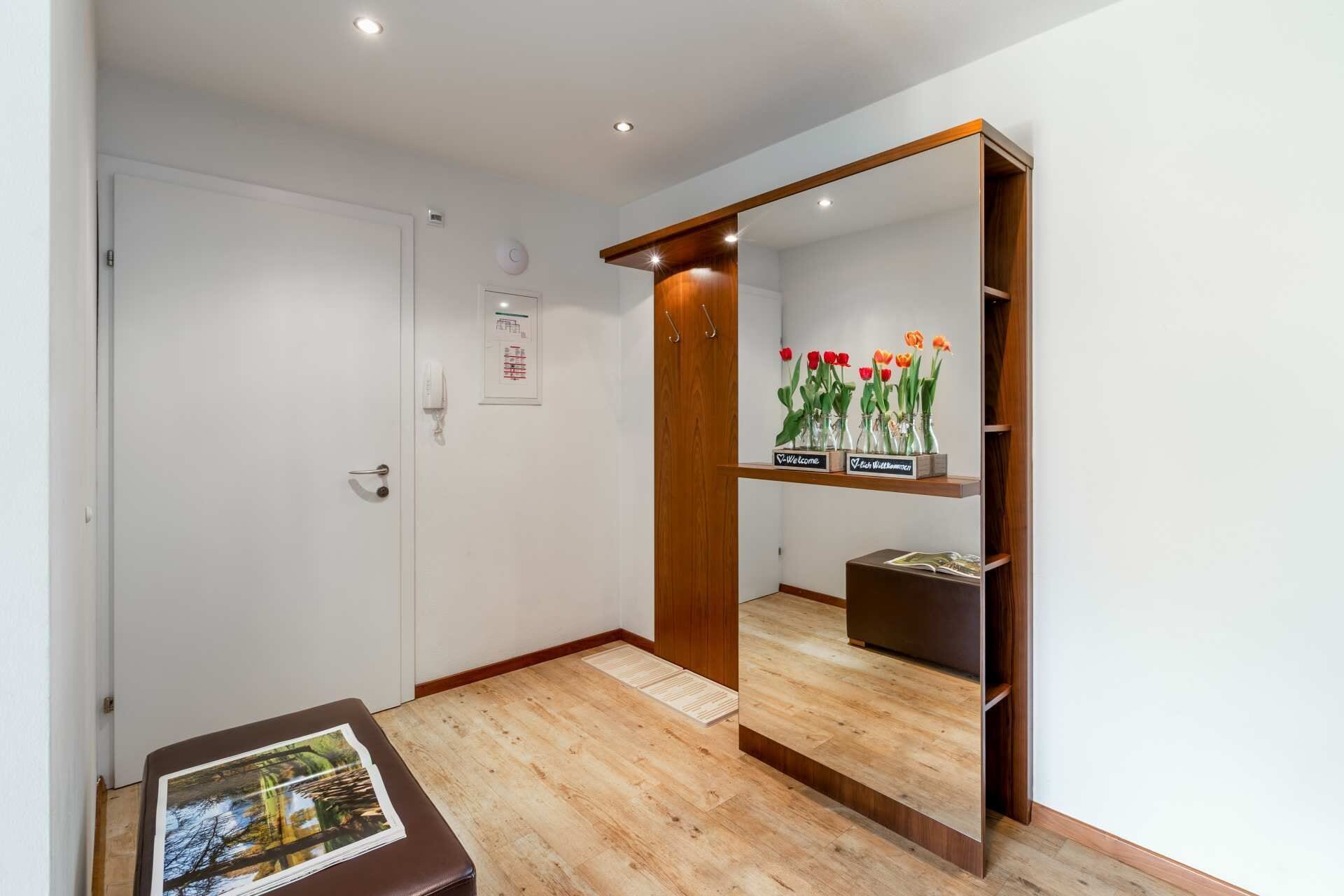 Une chambre d’hôtel moderne avec un plancher en bois, un grand miroir, une pancarte de bienvenue et une petite table sur laquelle est posée une photo.