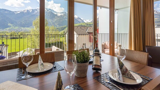 Mesa para comer preparada con copas de vino, platos y una botella, con vistas a un paisaje montañoso y un balcón.