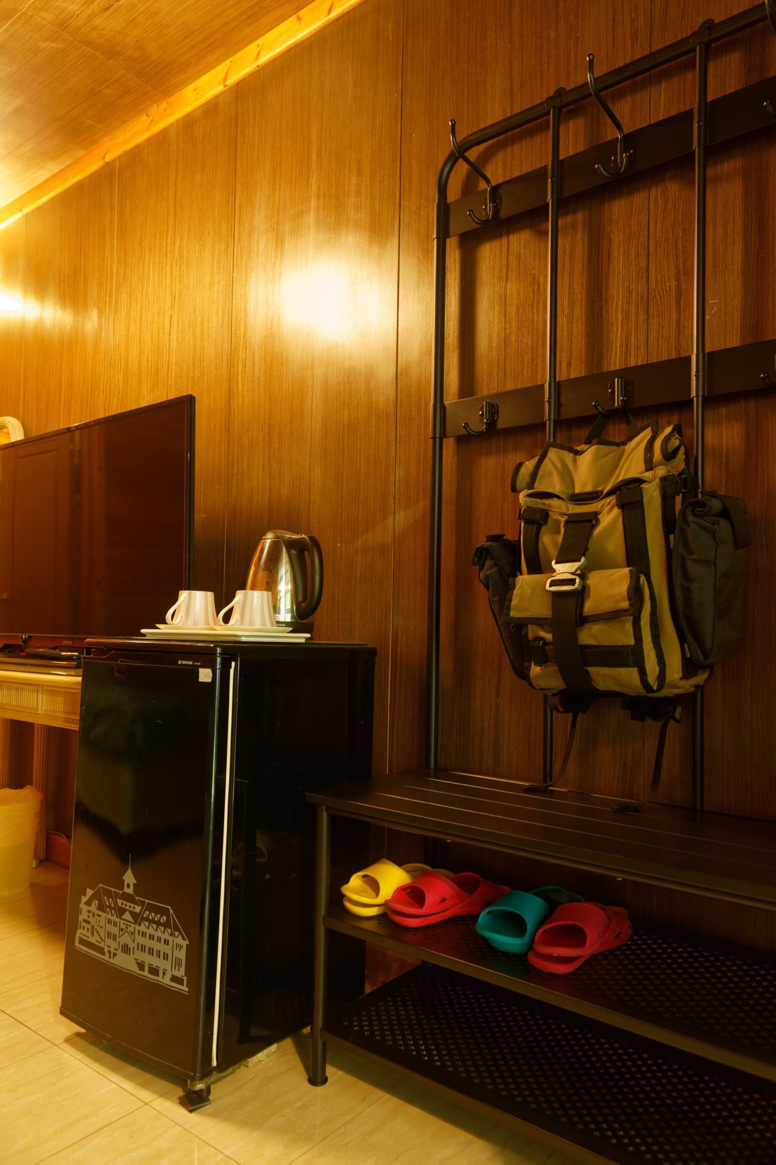Una habitación compacta con pared de madera, un refrigerador negro pequeño, cafetera, perchero para equipaje y zapatero con pantuflas de colores.