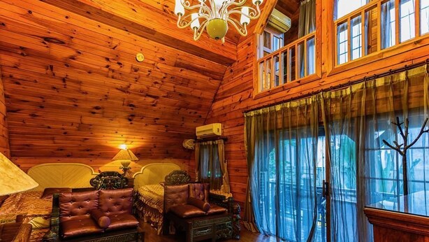 Interior de una cabaña de troncos con techo de madera, una araña, una cama con dosel y un amplio ventanal con cortinas.