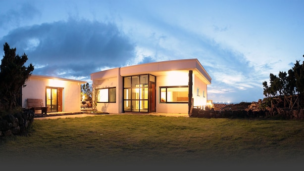 A modern two-story house with large glass windows and doors, a well-maintained lawn, and a stone wall in the background. - Void (Jeju City)