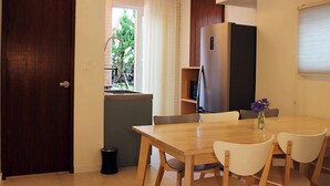A compact kitchen with a wooden table and chairs, a refrigerator, and a window with curtains. - Void (Jeju City)