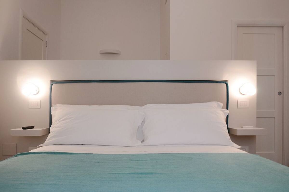 Cama bem arrumada com roupa de cama branca e um cobertor azul-esverdeado, acompanhada por mesas de cabeceira com luminárias.