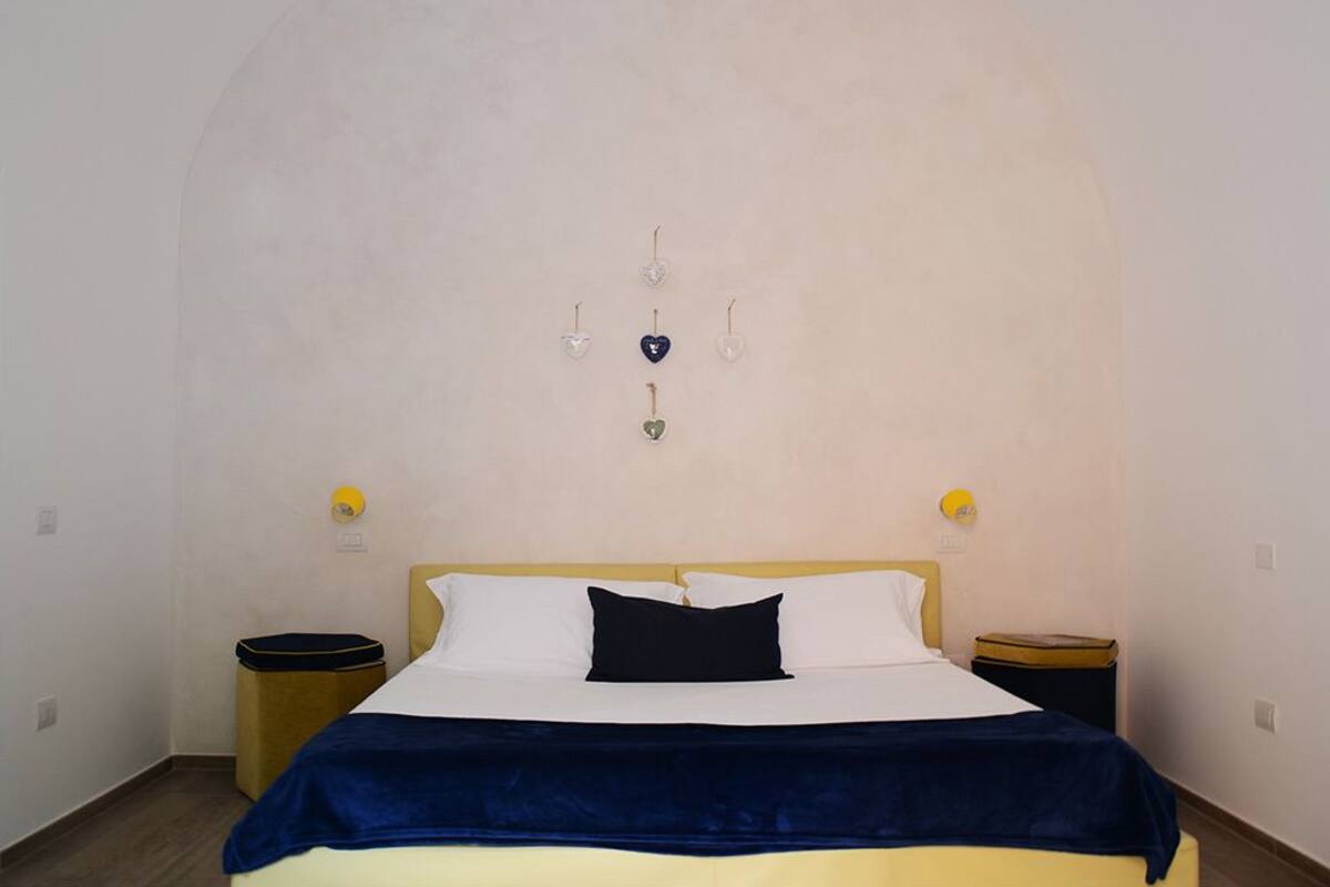 Quarto com cama, mesa de cabeceira, lixeira e luminária na parede.