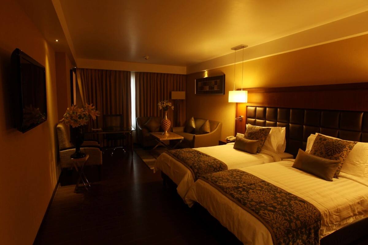 Quarto de hotel com duas camas, uma escrivaninha, uma cadeira e uma luminária.