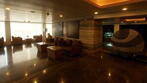Un hall espacioso de diseño moderno, con un amplio espacio de estar con sofás y una mesa de centro, una escultura decorativa única y un gran ventanal que proporciona luz natural.