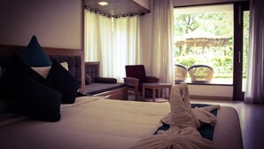 A hotel room with a bed, a chair, a small table, and a view of greenery outside. - Namami Ganges Resort (Narendranagar)