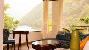 A room with a view of a forested hillside, a round glass table, a sofa, and a chair. - Namami Ganges Resort (Narendranagar)