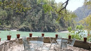 An outdoor seating area with tables and chairs overlooking a river, surrounded by trees and a mountainous backdrop. - Namami Ganges Resort (Narendranagar)