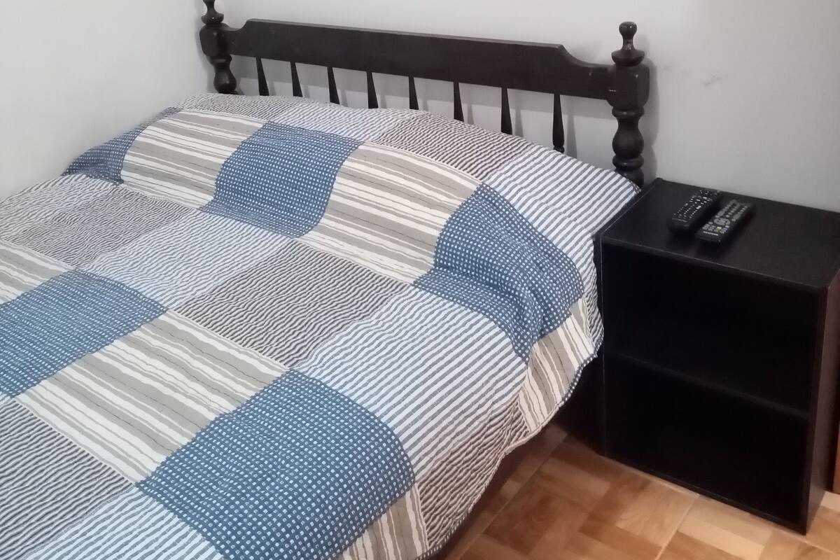 Een bed met een gestreept en bewerkt beddegoed, een houten hoofdbord, een nachtkastje met een afstandsbediening, en een houten vloer.