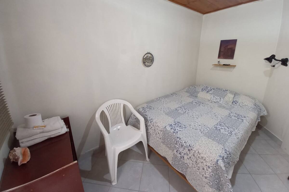 Een kleine, schone kamer met een bed, een witte plastic stoel, een klein tafeltje met een handdoek en een rol toiletpapier, en een wandplank met een foto.