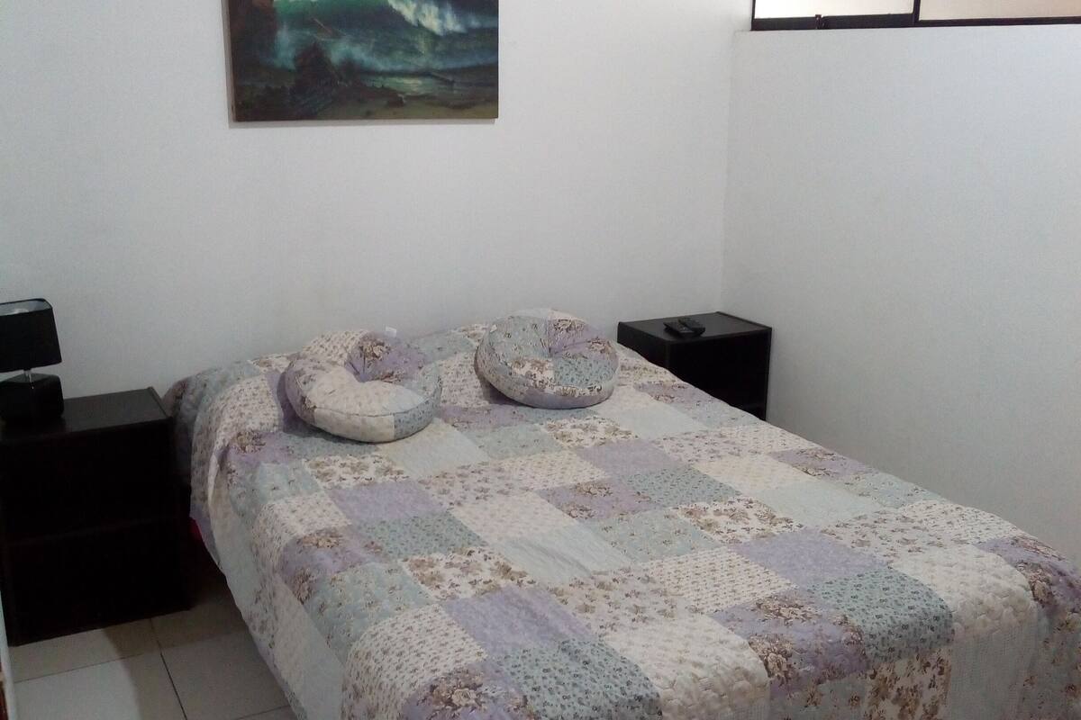 Een slaapkamer met een bed, twee nachtkastjes, een aan de muur hangend schilderij en een raam met jaloezieën.