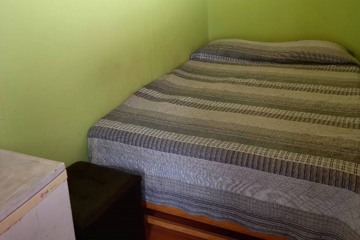 Een kleine kamer met een enkel bed, een groene wand, een houten plafond, een bed met een gestreept dekbed, een klein tafeltje en een ventilator.