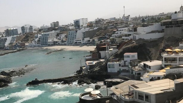 A coastal cityscape with buildings along the beach, rocky shoreline, and a clear view of the sea.