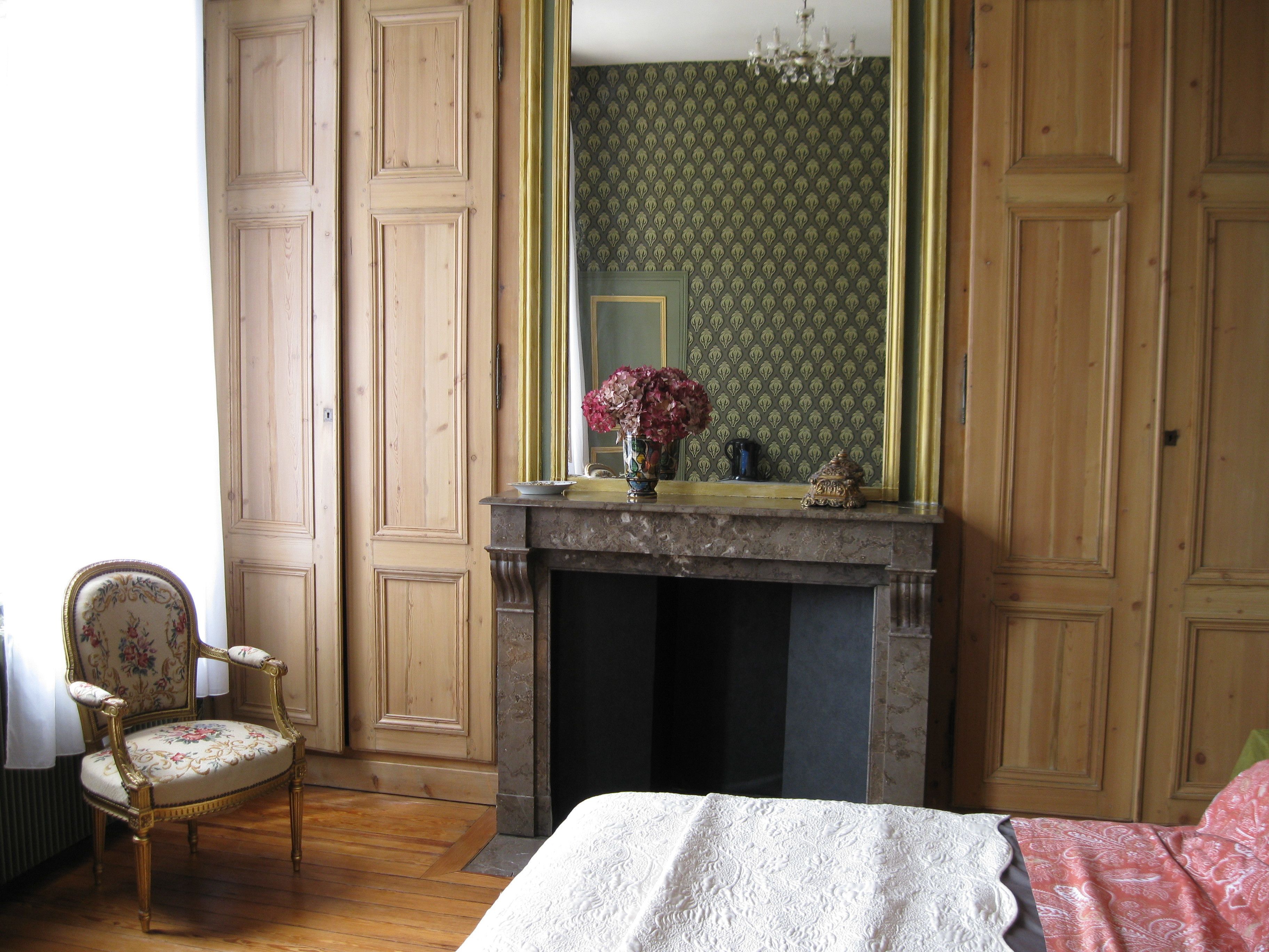Une pièce dotée d’un plancher en bois, d’un lit recouvert d’une couette blanche, d’une chaise à l’assise à motifs floraux, d’une cheminée ornée d’un vase de fleurs et d’un miroir placé devant un papier peint à motifs verts.