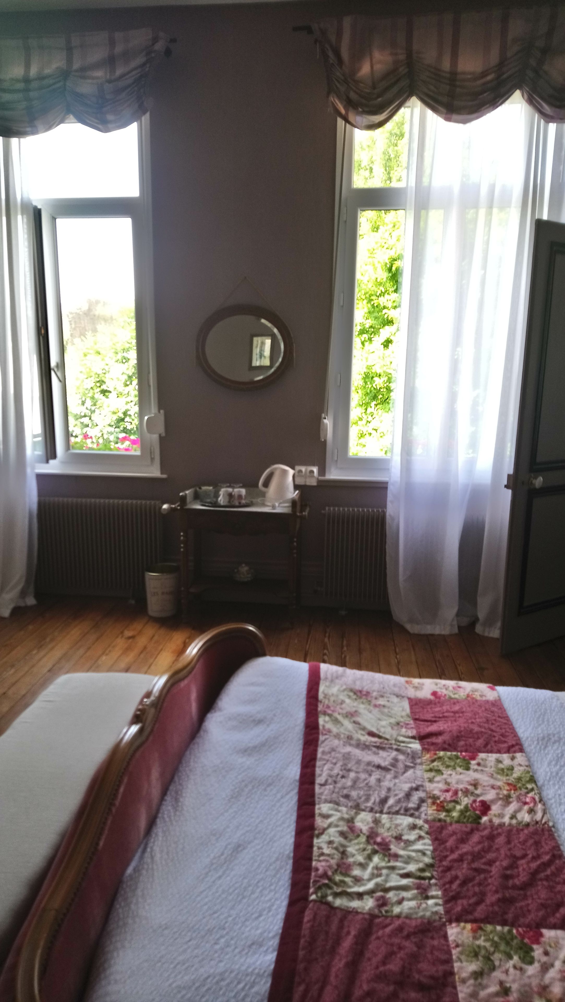 Een slaapkamer met een bed, nachtkastje, spiegel, radiator en een raam met gordijnen.