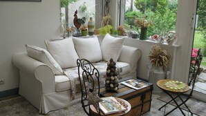 A cozy living room with a white sofa, a coffee table with a book, and a view of greenery outside. - Maison d'Hôtes Cap et Marais d'Opale (Saint-Omer)