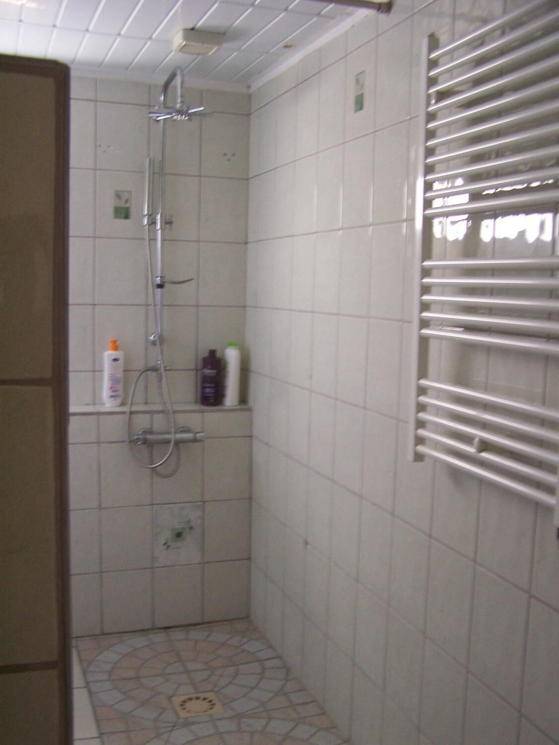 A small, tiled shower area with a handheld showerhead, a wall-mounted towel rack, and a few toiletries on a shelf.