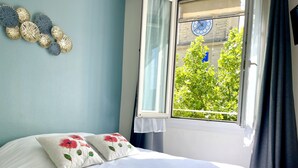 Un lit bien fait, agrémenté d’oreillers à motifs floraux, une fenêtre donnant sur un paysage verdoyant, une horloge et des œuvres d’art décoratives accrochées au mur.