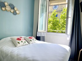 Un lit bien fait, agrémenté d’oreillers à motifs floraux, une fenêtre donnant sur un paysage verdoyant, une horloge et des œuvres d’art décoratives accrochées au mur.