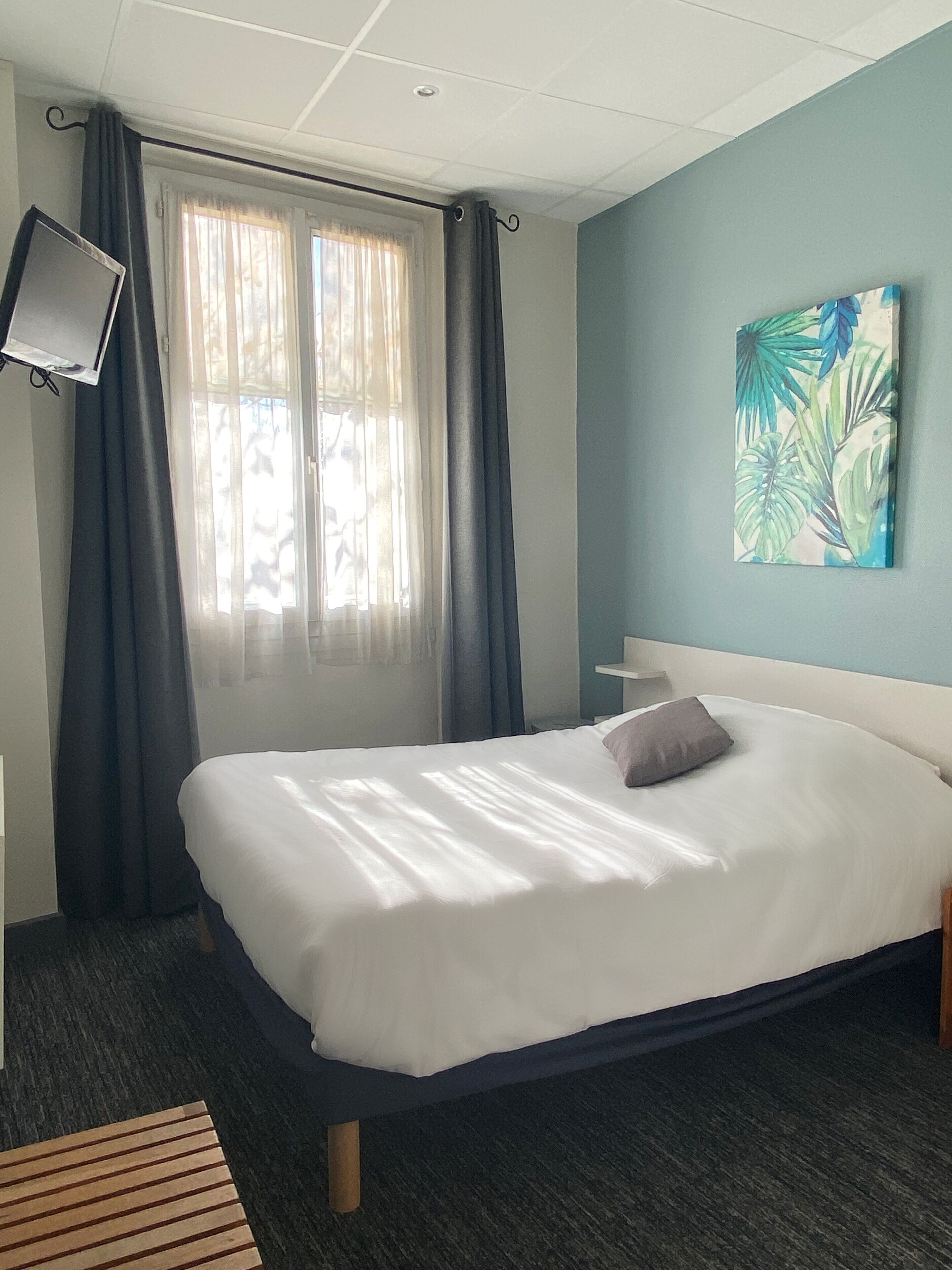 A hotel room with a bed, a flat-screen TV mounted on the wall, a painting of tropical leaves, and a window with curtains.