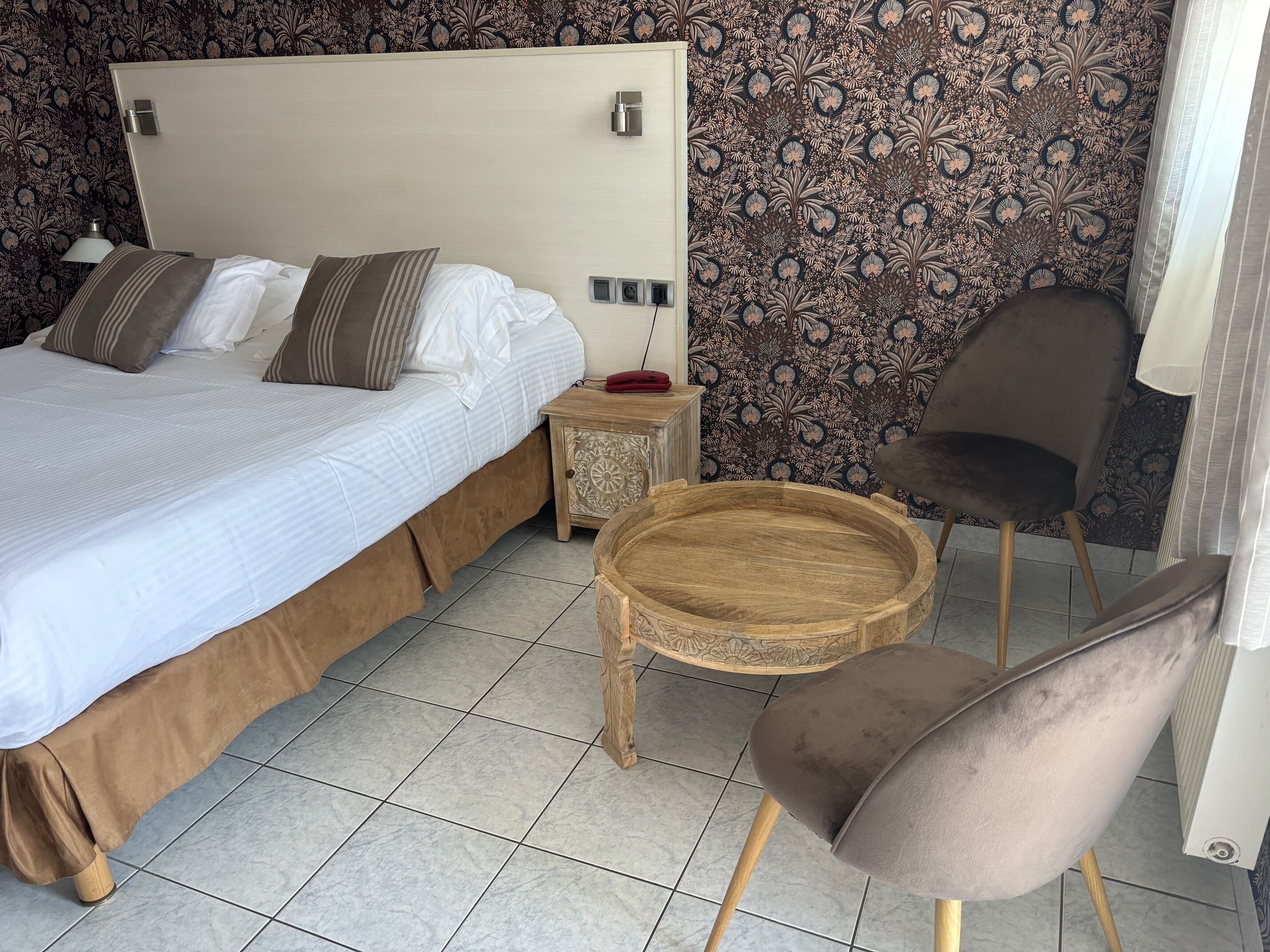 A neatly made bed with a wooden headboard, a bedside table with a lamp, a round wooden tray, and a chair.