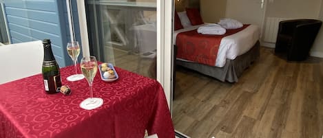 A hotel room with a bed, a small table with a bottle and glasses, and a view of the outside.