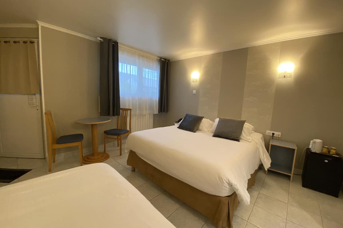 Ein Hotelzimmer mit einem großen Bett, einem kleineren Bett, einem Sessel, einem Tisch und einem Nachttisch mit einer Lampe.