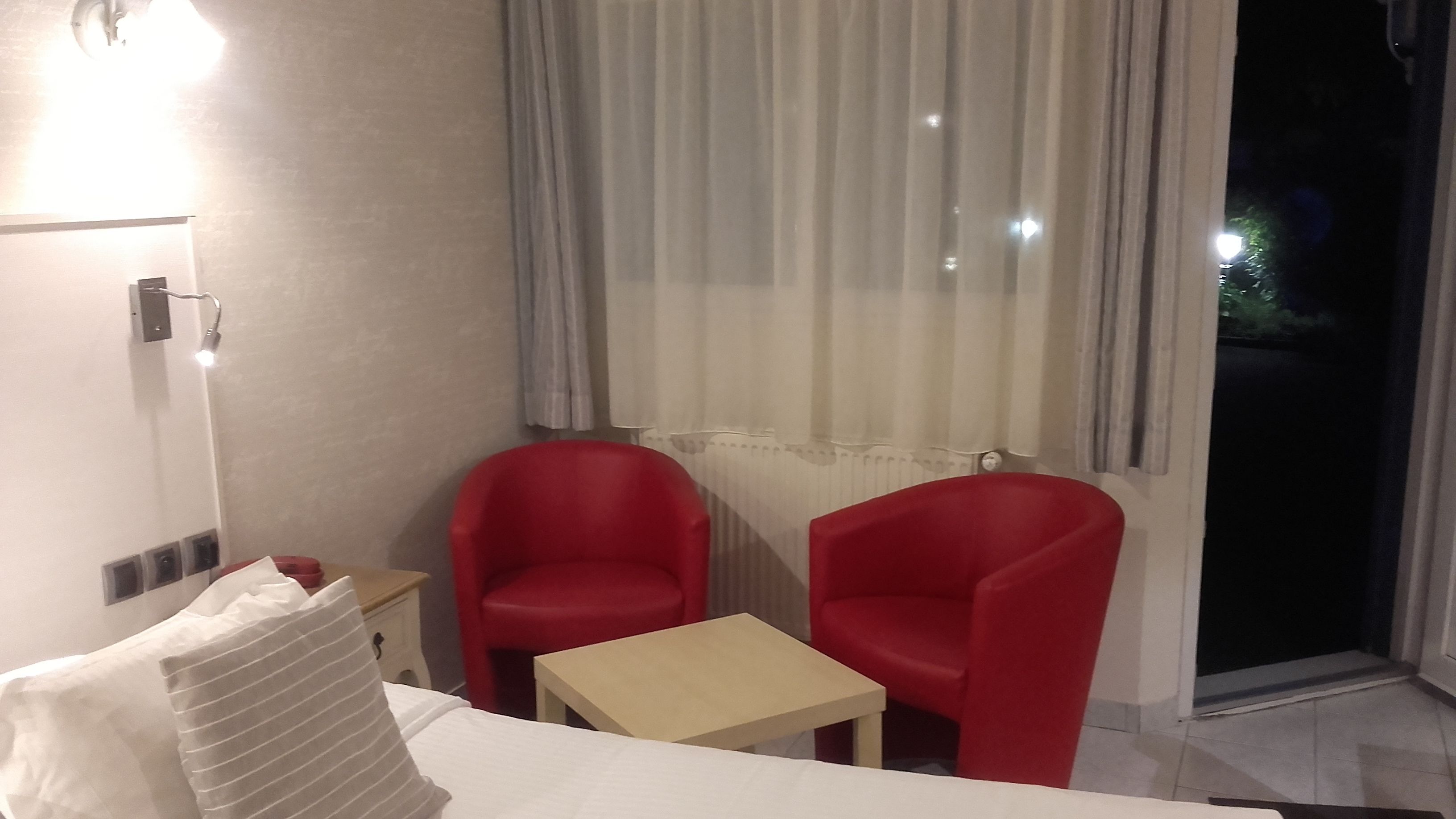 Une chambre d’hôtel avec un lit, deux chaises rouges, une petite table, une fenêtre avec des rideaux et un radiateur.