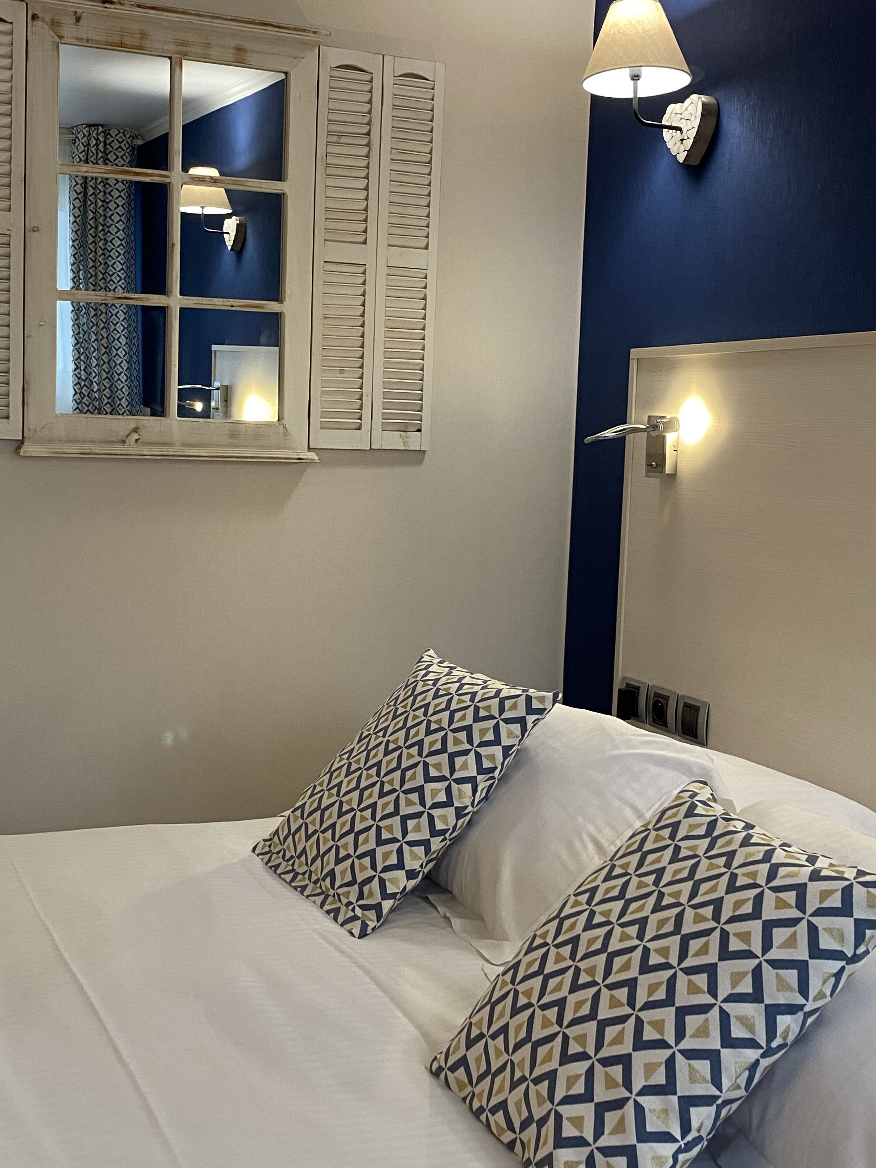 Un lit avec des coussins à motifs, une fenêtre avec des volets et une lampe fixée au mur.