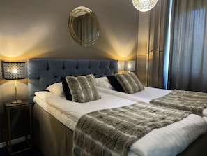 A neatly made double bed with a tufted headboard, two bedside lamps, a round mirror on the wall, and a view of a window with curtains. - Golden Circle Suites (Rovaniemi)