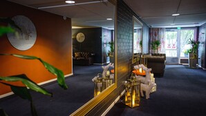 Un vestíbulo con un espejo grande que refleja un reloj, una zona de estar con un sofá y sillones, y un mostrador de recepción con una planta.