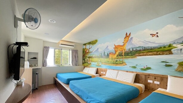 A hotel room with two beds, a mural of a forest scene, a fan, and a desk with a computer.