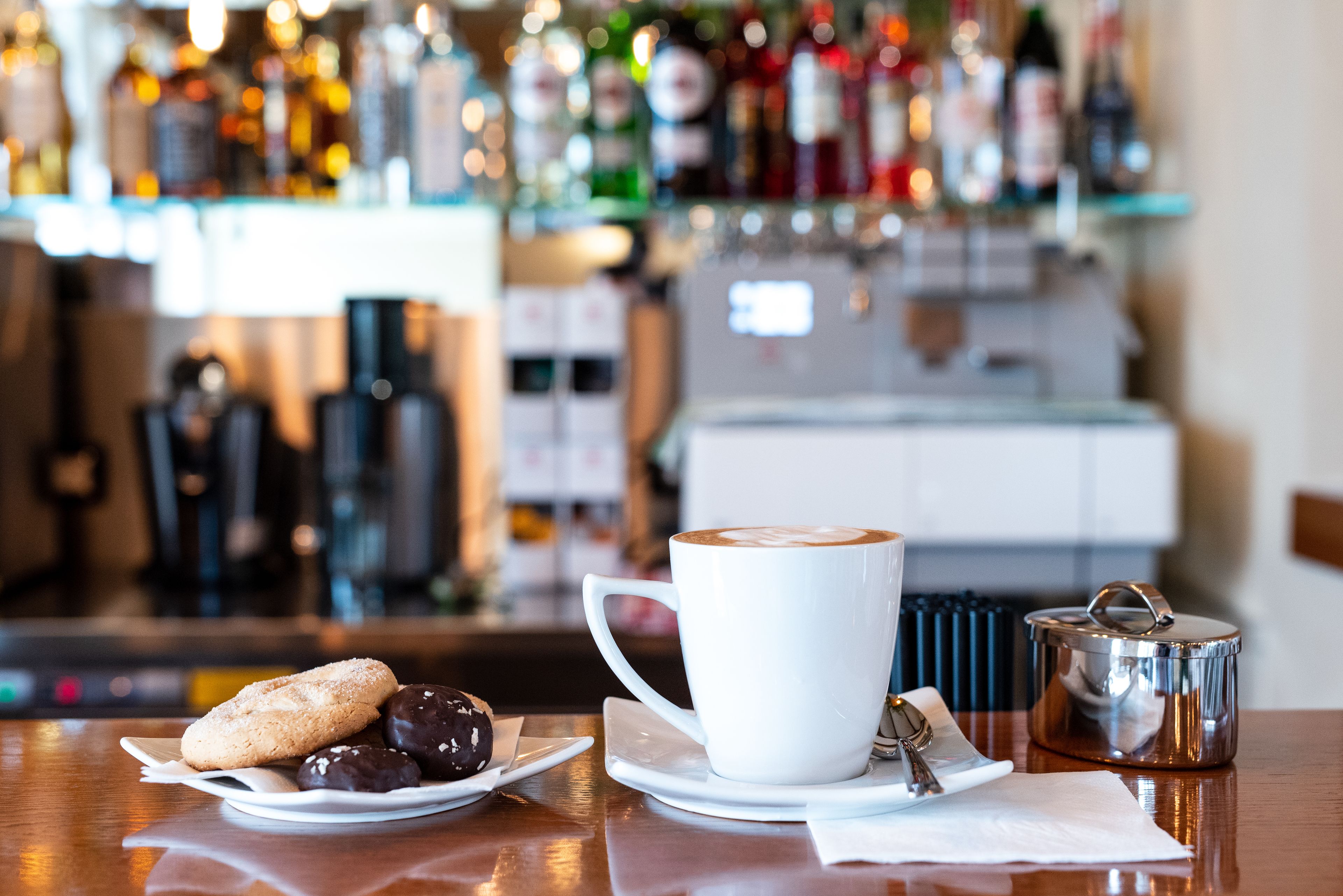 Un ambiente de bar con una taza de café blanco, un plato de galletas y un termo sobre una mesa de madera.
