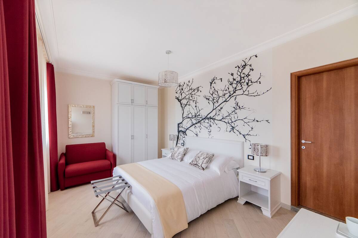 A bedroom with a bed, a red chair, a wardrobe, a nightstand, and a wall art piece.