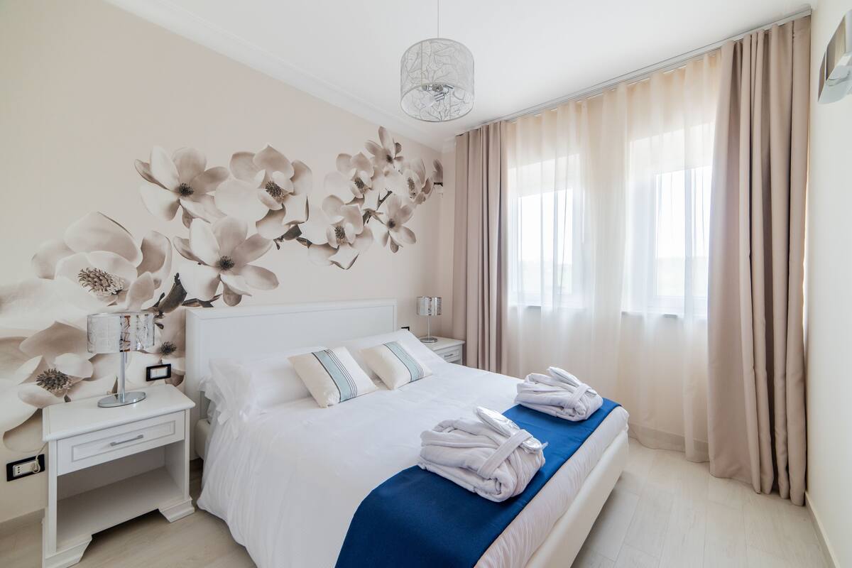 A hotel room with a large bed, a nightstand, a wall-mounted lamp, and a window with curtains.