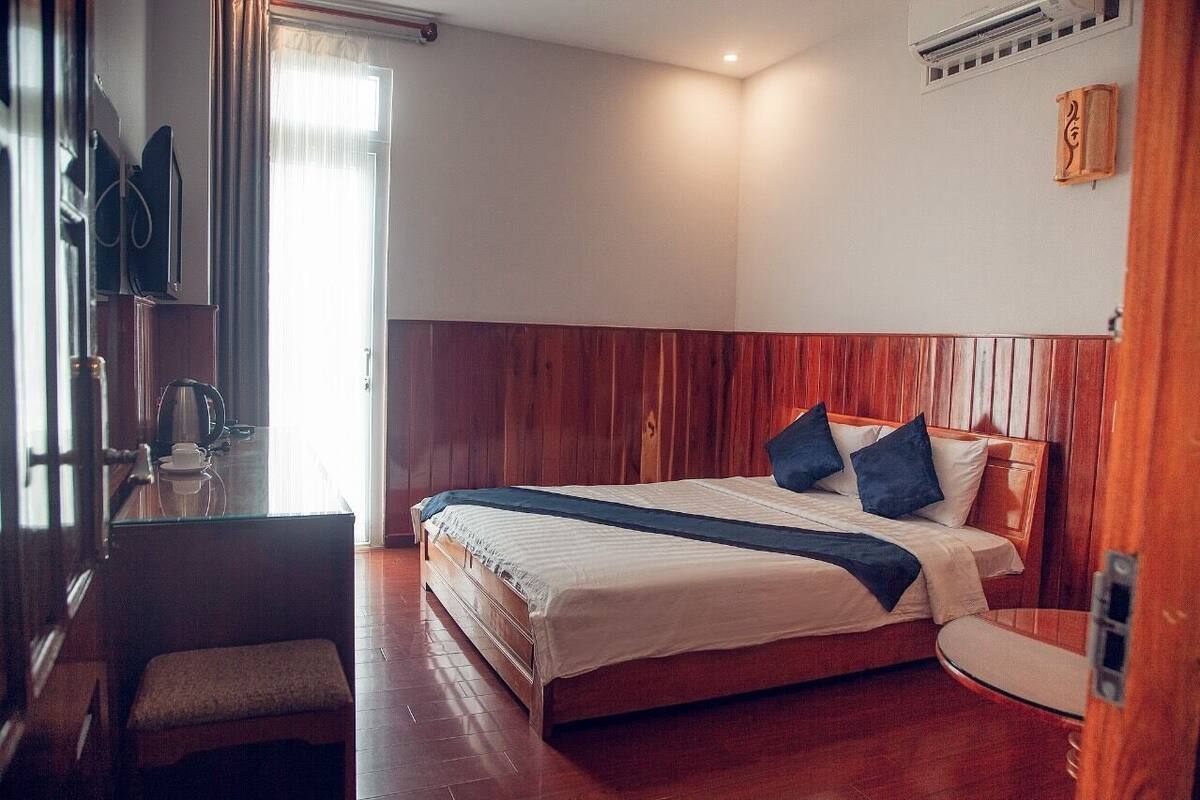Une chambre d’hôtel avec une tête de lit en bois, un lit recouvert de draps blancs et agrémenté d’oreillers foncés, une petite table de chevet avec une tasse et un livre, et une fenêtre avec des rideaux.