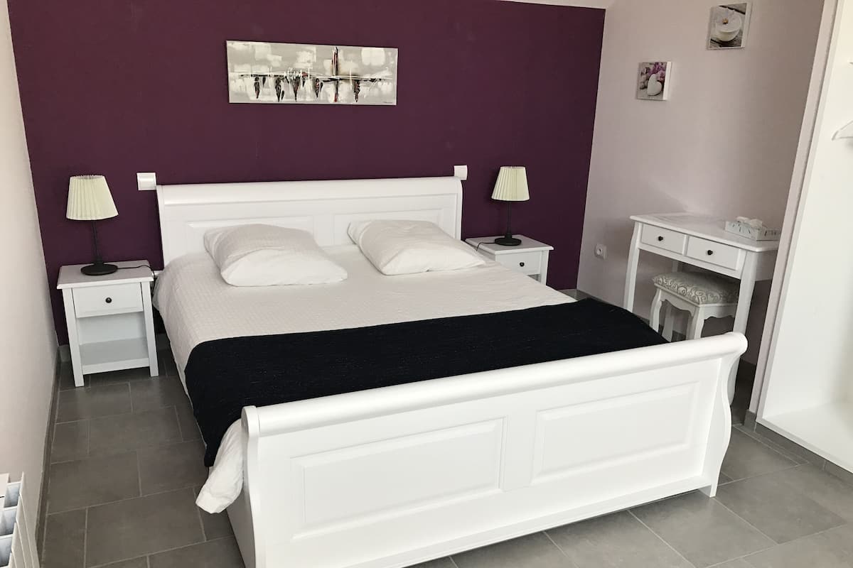 Un lit bien fait, avec du linge de lit blanc, un plaid noir et une tête de lit blanche. On y trouve deux tables de chevet avec des lampes, une commode et une petite commode blanche avec un miroir.