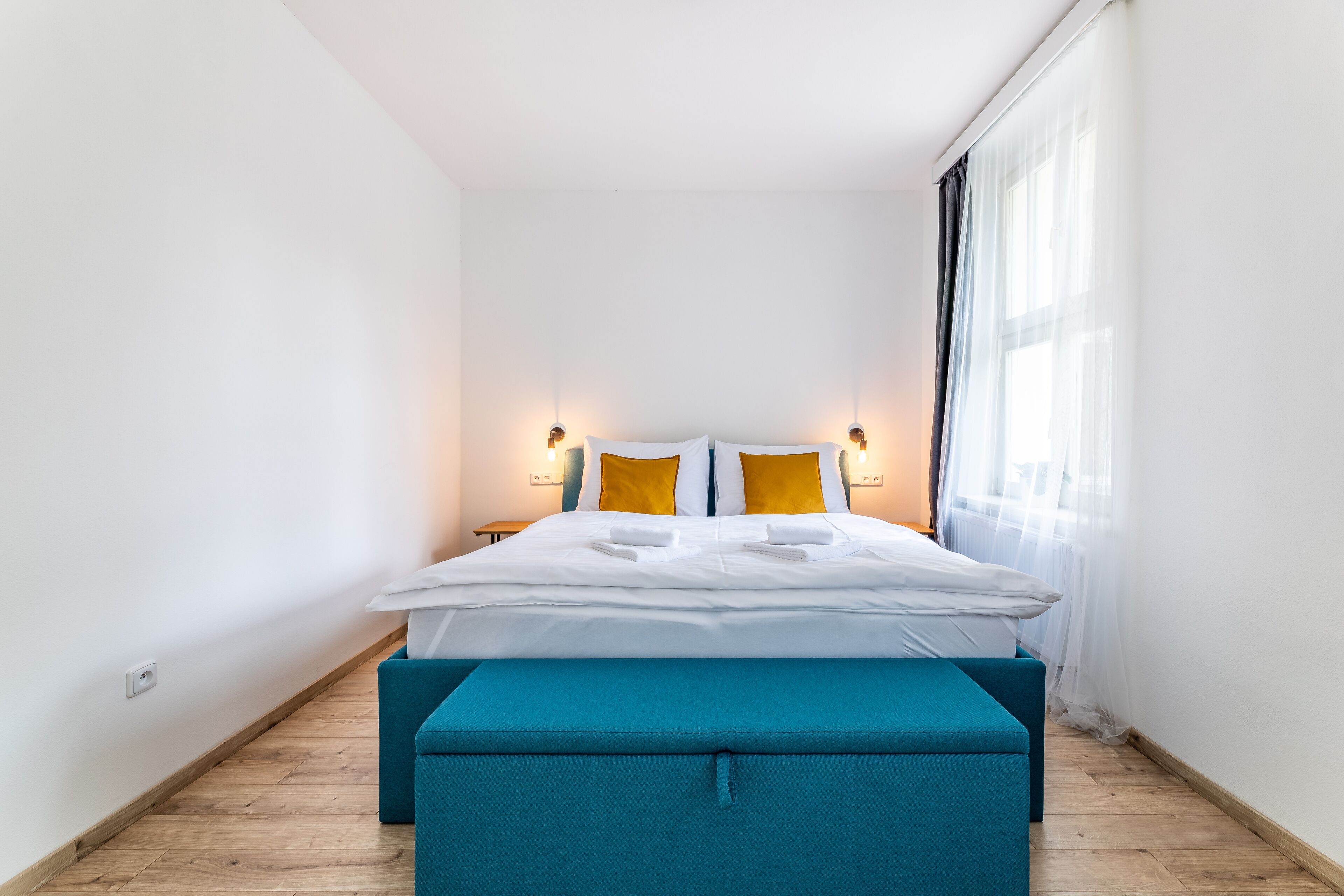 A modern hotel room with a neatly made bed, two bedside tables, and a large window with sheer curtains.