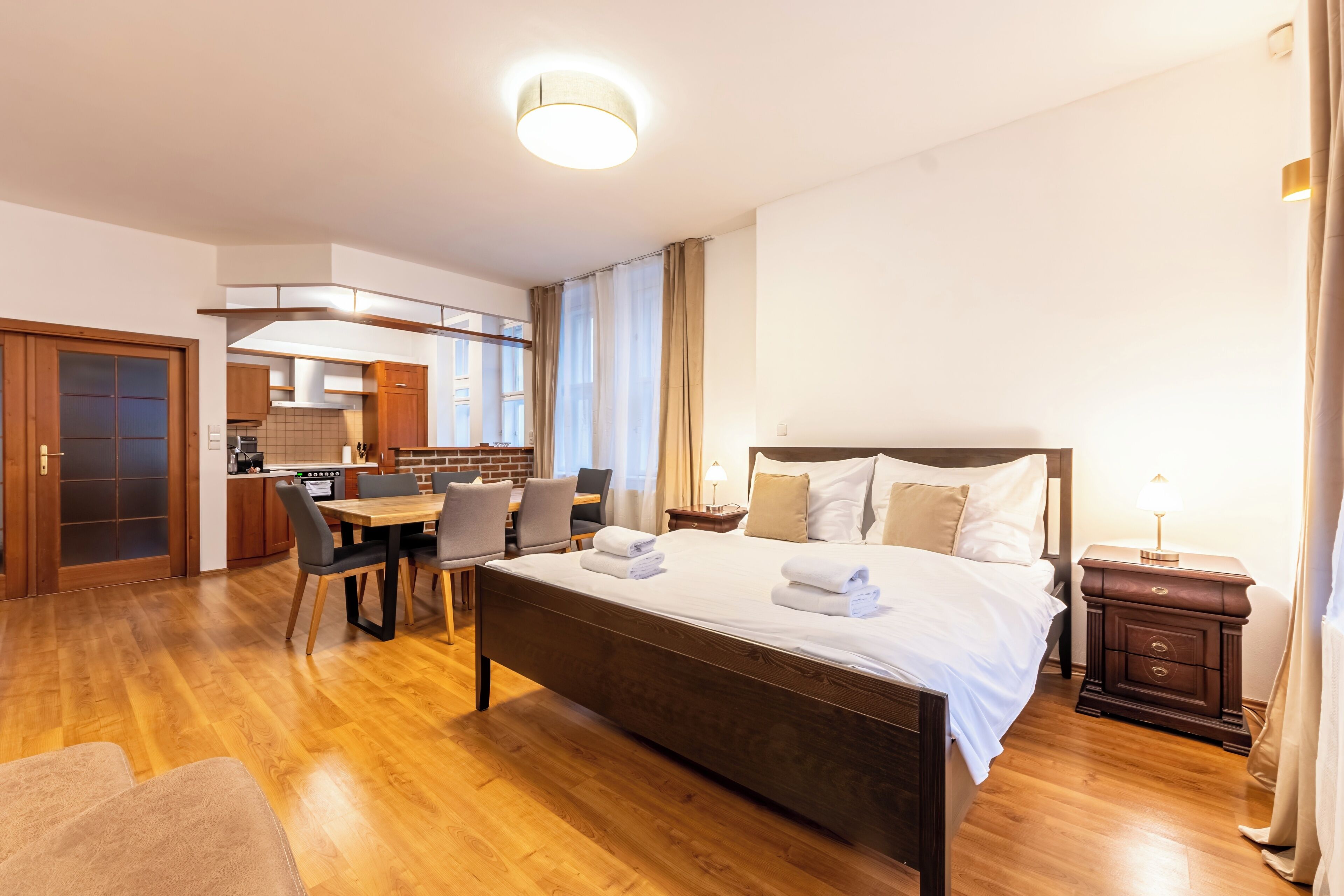 Habitación de hotel con piso de madera, cama con ropa blanca, mesa de comedor con sillas, zona de cocina y baño.