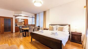 Habitación de hotel con piso de madera, cama con ropa blanca, mesa de comedor con sillas, zona de cocina y baño.