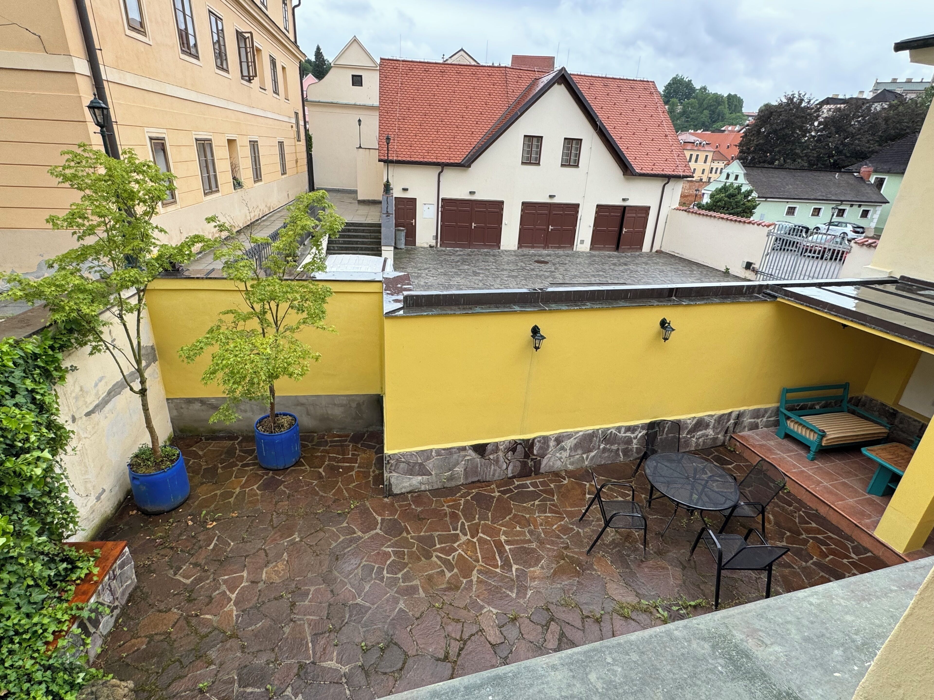 A courtyard with stone flooring, a yellow wall, a small tree, and a table with chairs.