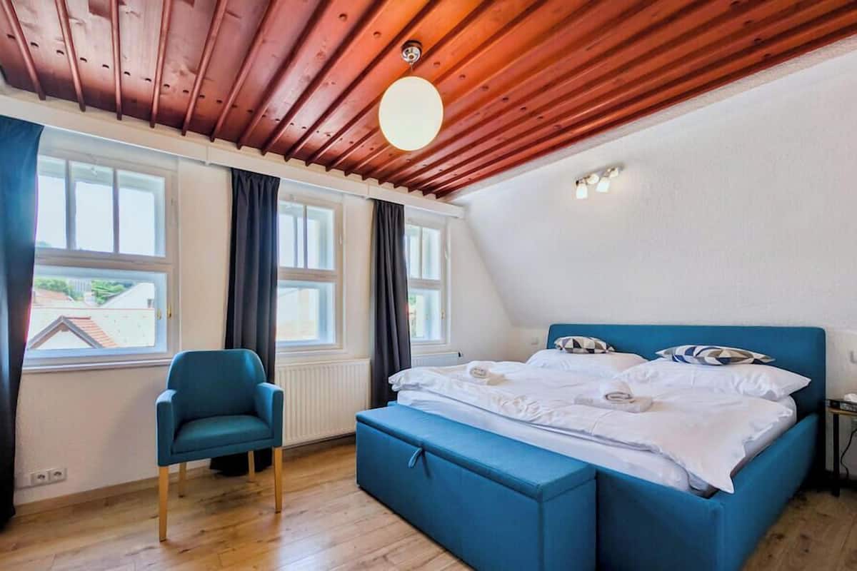 A bedroom with a wooden ceiling, a bed with blue bedding, a chair, and large windows.