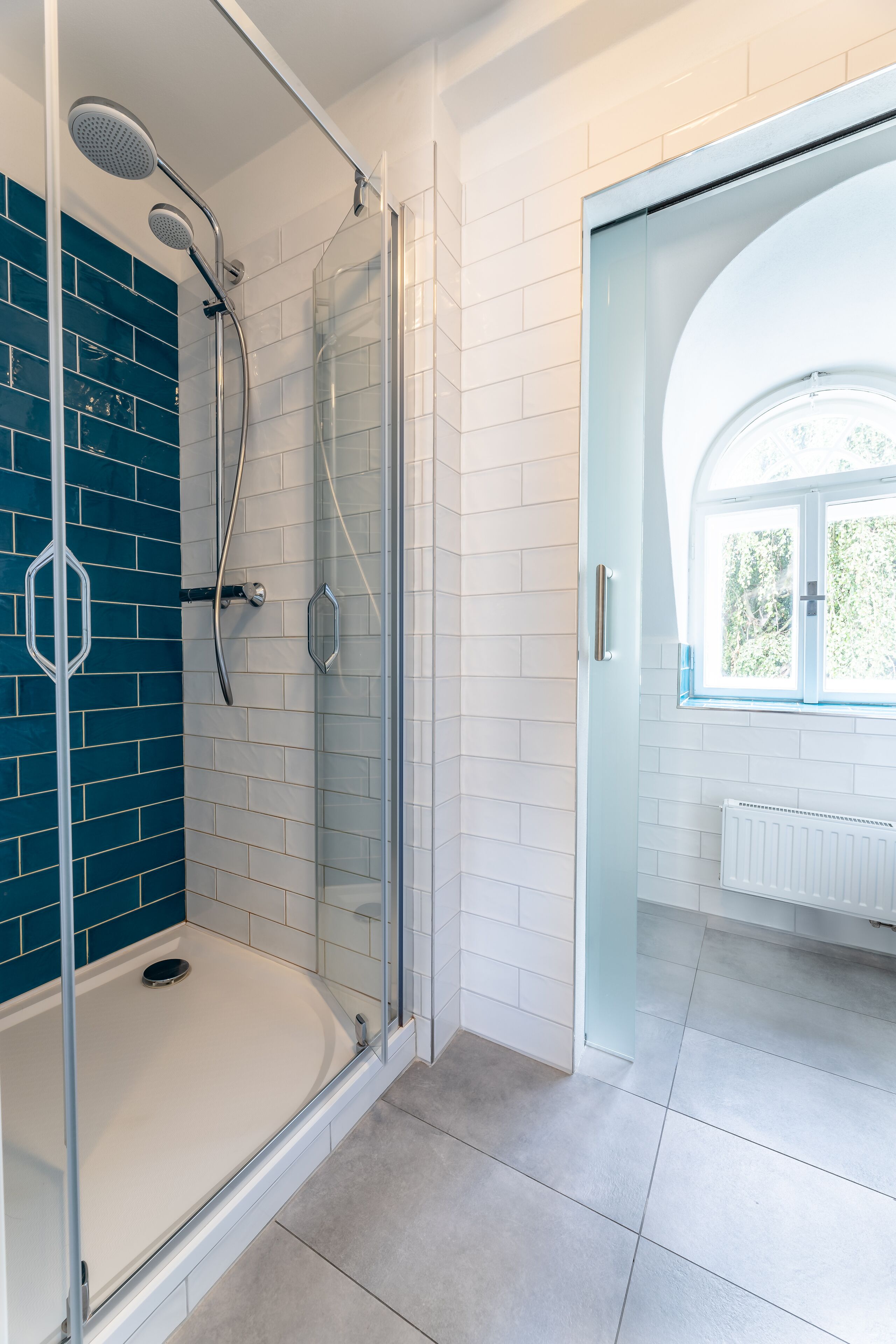 A modern bathroom with a glass-enclosed shower, white tiled walls, and a view of the outdoors through a window.