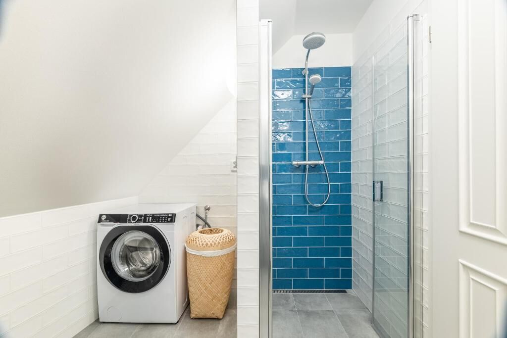 Un área de lavandería con lavadora y cesta, una ducha con pared de azulejos azules y puerta de vidrio.
