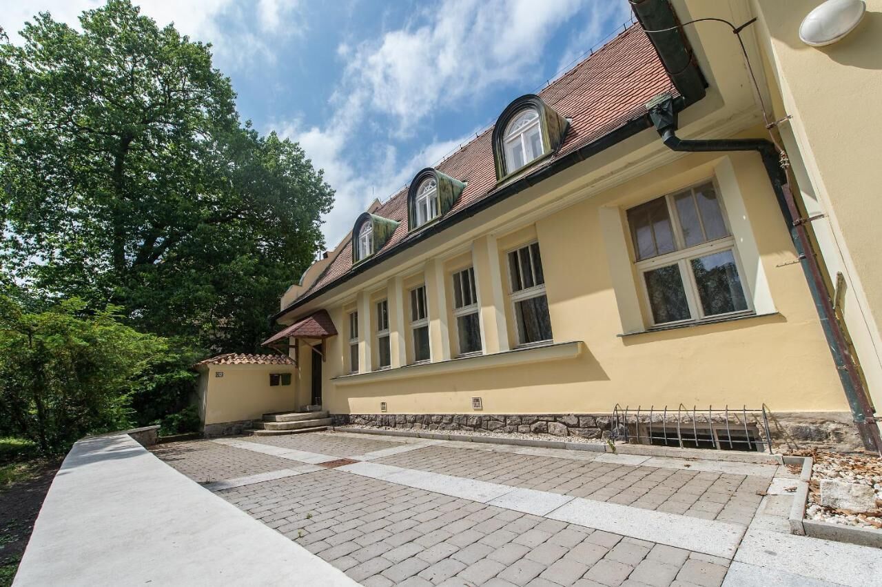 A building with a yellow facade, multiple windows, a tiled roof, and a paved entrance area.